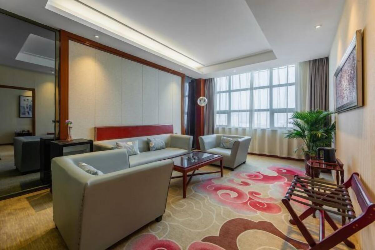 A modern hotel room with a sofa set, a coffee table, a wooden bench, and a large window providing natural light.