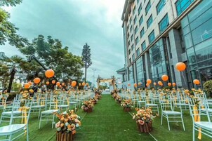 Un espacio para eventos al aire libre con hileras de sillas y mesas blancas, decorado con esferas naranjas y arreglos florales.