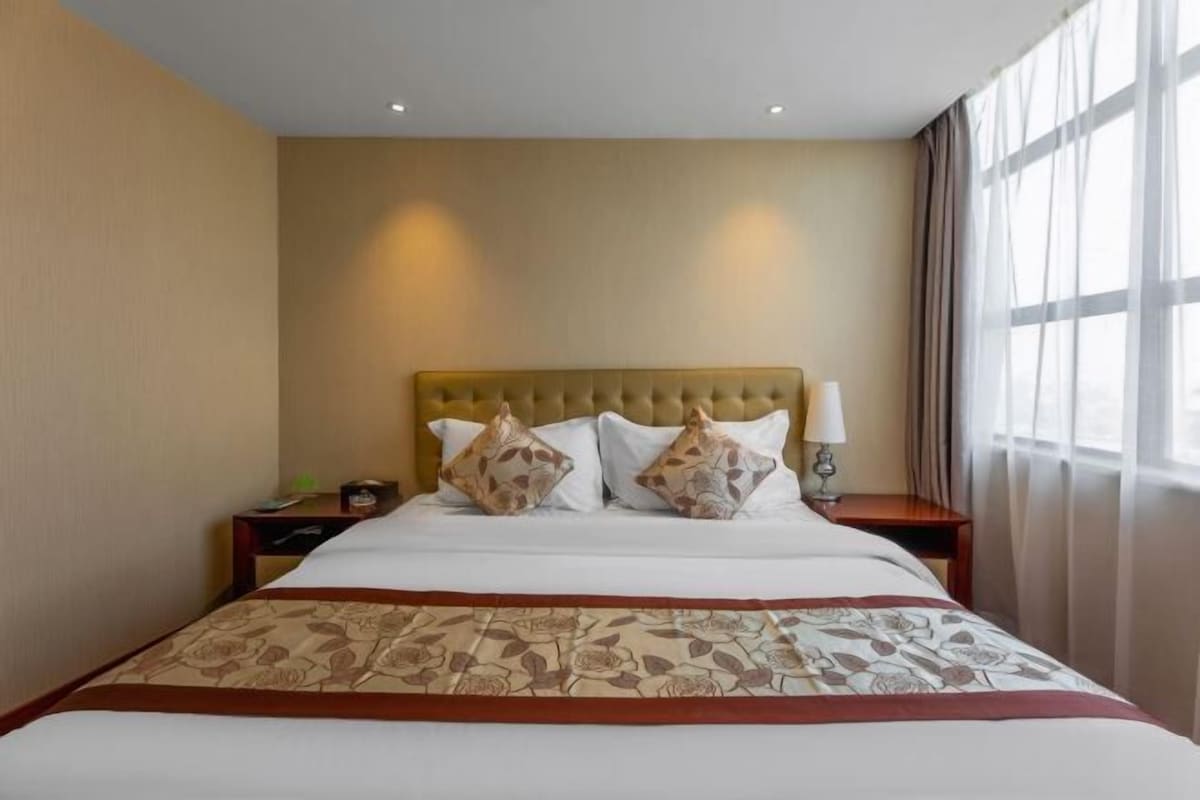 Una habitación de hotel con una cama grande, un cabecero, dos mesitas de noche y una ventana con cortinas.