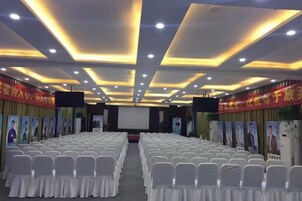 Un salón de conferencias con filas de sillas blancas, un escenario y una pantalla.