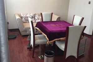 Een eethoek met een tafel bedekt met een paarse tafelkleed, omringd door zes stoelen. Een prullenbak is op de vloer te zien.