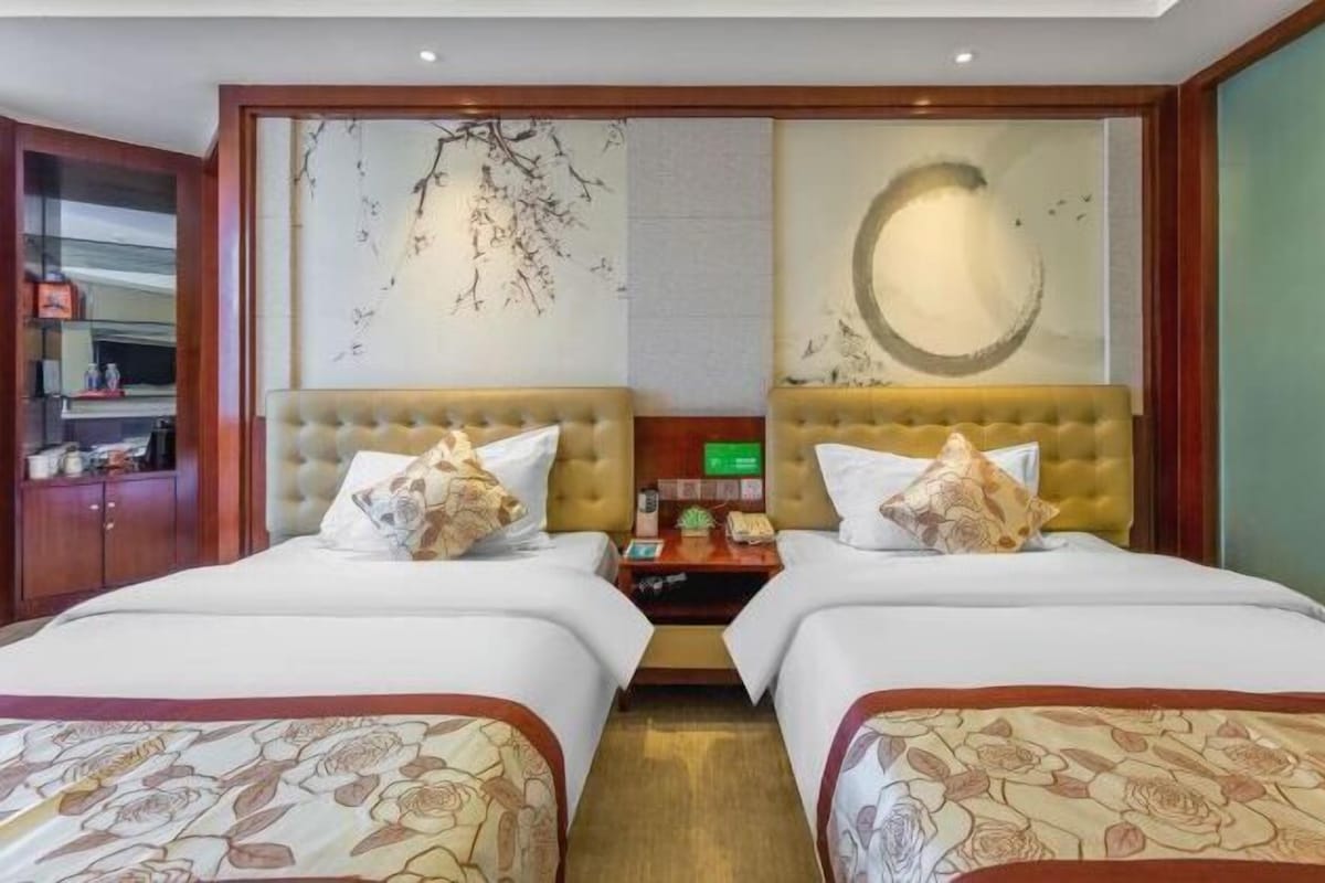 Habitación de hotel con dos camas, un cabecero de madera, una pared con un diseño circular y una mesita de noche pequeña con una lámpara.