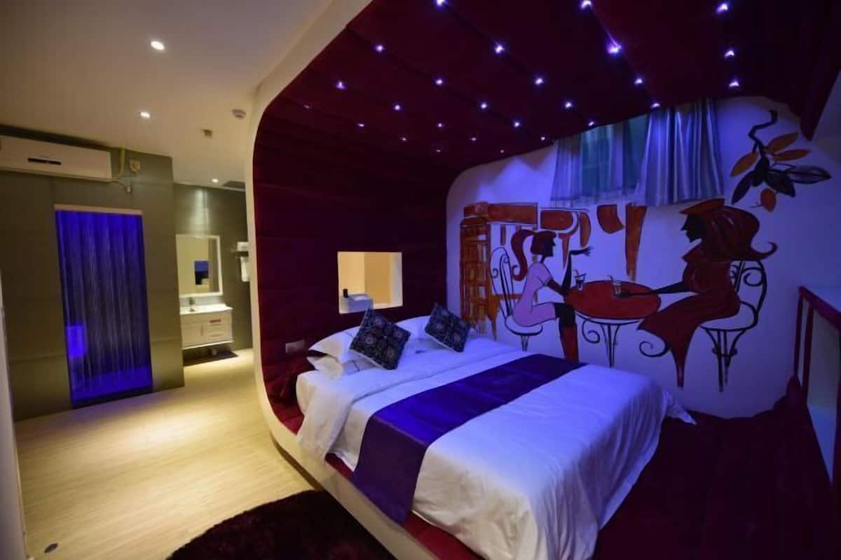 A modern bedroom with a curved ceiling, a bed with a purple accent, a mural, and a bathroom with a blue-lit shower.