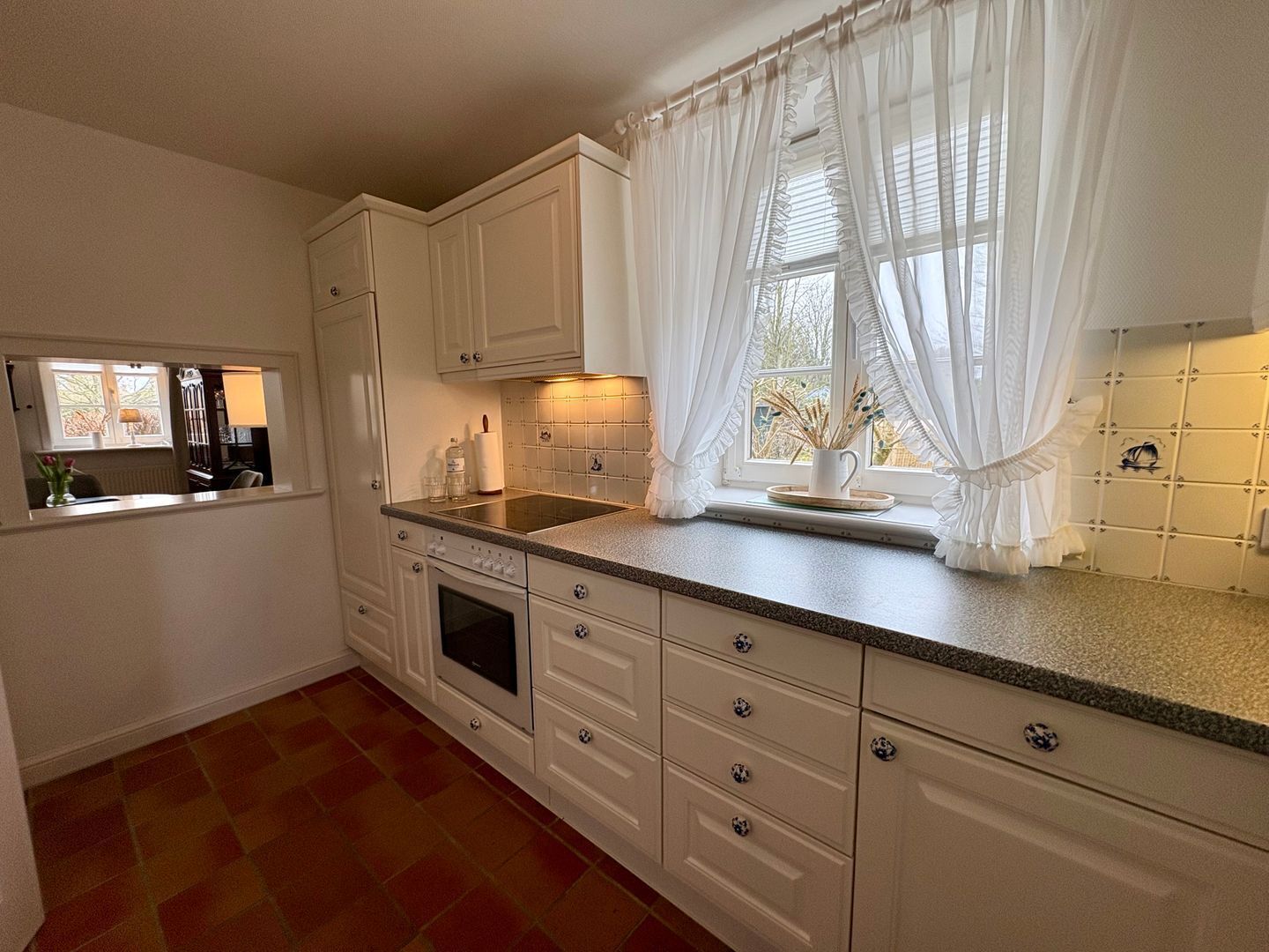 Una cocina con armarios blancos, estufa, lavamanos y ventana con cortinas ligeras.