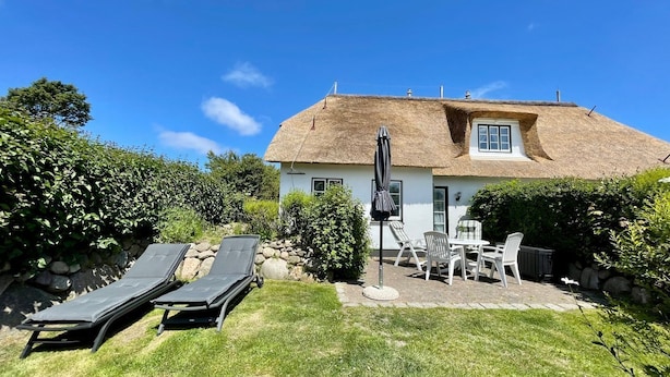 A house with a thatched roof, a stone wall, and a lawn with two lounge chairs.