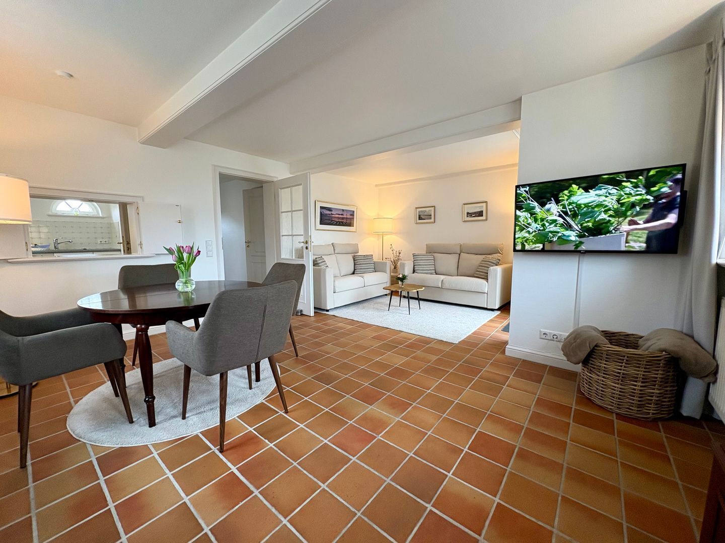 A modern living room with a dining area, a TV mounted on the wall, and a kitchen visible through an open door.