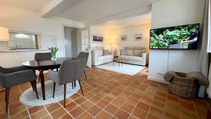 A modern living room with a dining area, a TV mounted on the wall, and a kitchen visible through an open door.