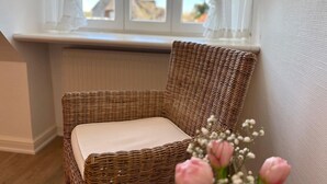 Ein Korbstuhl mit Kissen, ein Fenster mit weißen Vorhängen und eine Vase mit Blumen.