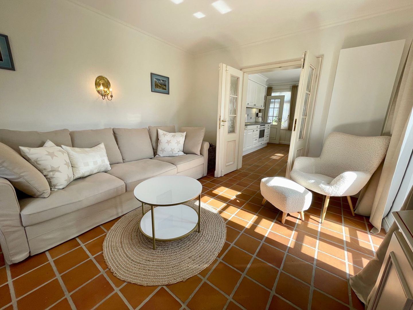 Un salon avec un canapé beige, une table basse ronde et un fauteuil blanc. On aperçoit une cuisine au fond.