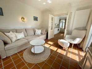 A living room with a beige sofa, a round coffee table, and a white armchair. A kitchen is visible in the background. - 3 Liv: 55 sqm on three levels offer plenty of space for up to 4 people - country house (Sylt)