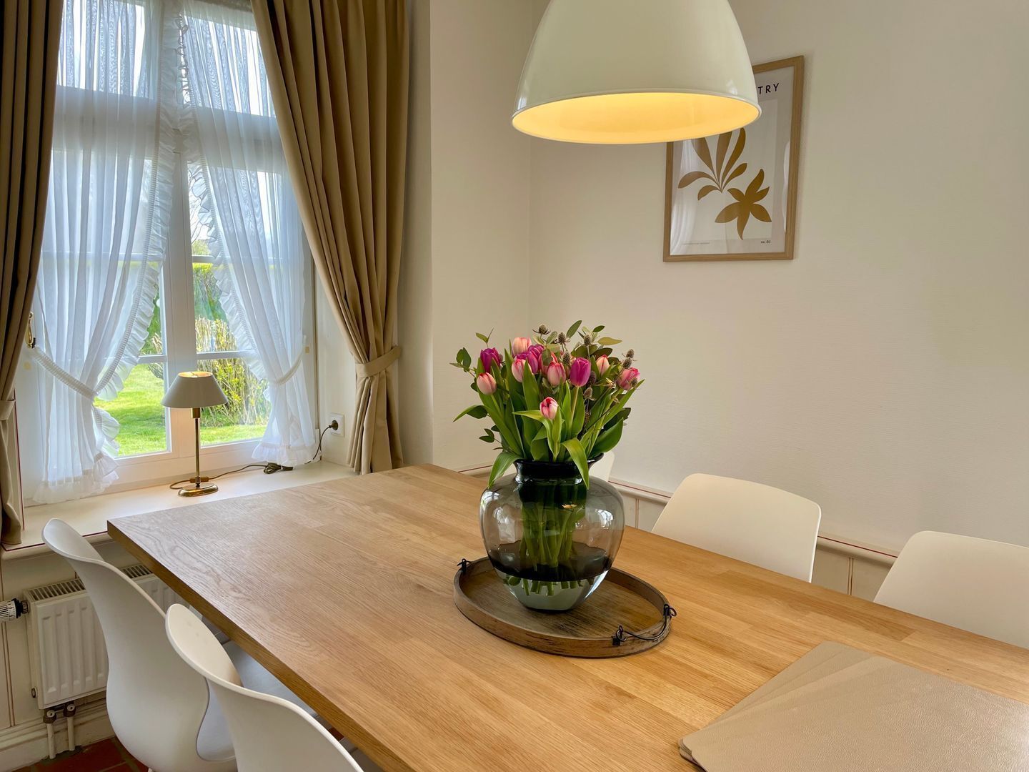 Ein Zimmer mit Holztisch, weißen Stühlen, einer Vase mit Blumen, einer Lampe und einem Fenster mit Vorhängen.