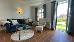 Ein Wohnzimmer mit blauem Sofa, rundem Couchtisch, weißem Sessel und Blick auf einen Garten durch große Fenster.