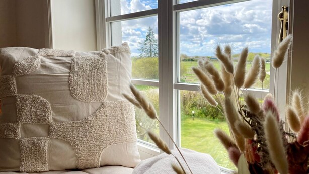Une pièce avec une grande fenêtre, un lit recouvert d’une couette blanche et beige, et un vase contenant des plantes séchées.