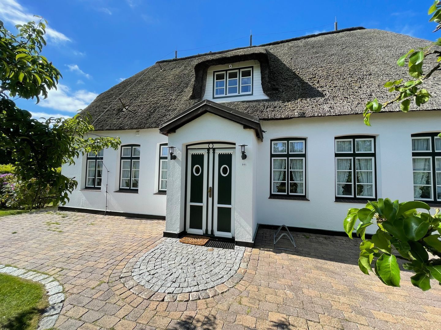 Een wit huis met een zwart dak, een cirkelvormige oprit en een kleine tuin.