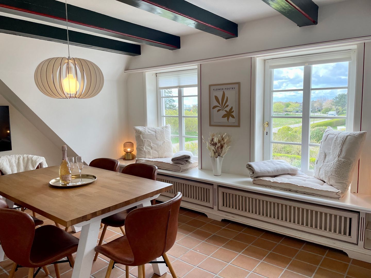Ein Essbereich mit Holztisch, Stühlen und einer Bank mit Handtüchern. Fenster bieten Blick auf Grünflächen.