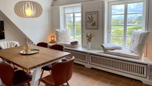 Un coin repas avec une table en bois, des chaises et un banc où sont posées des serviettes. Des fenêtres offrent une vue sur la verdure environnante.