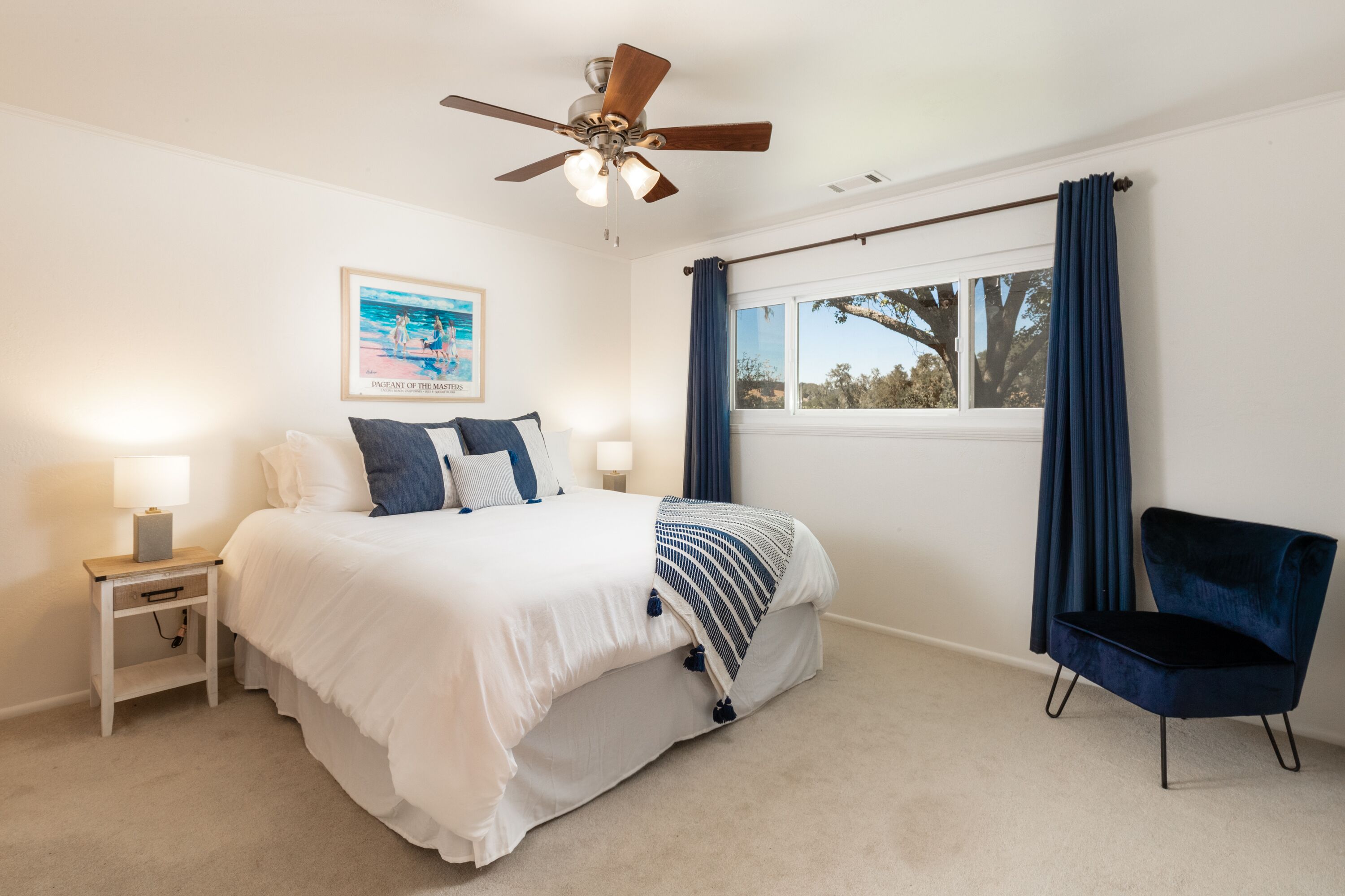 A bedroom with a bed, nightstands, a chair, a ceiling fan, a window with curtains, and a framed picture on the wall.