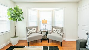 A room with two armchairs, a small table with a lamp, a large potted plant, and a bay window.