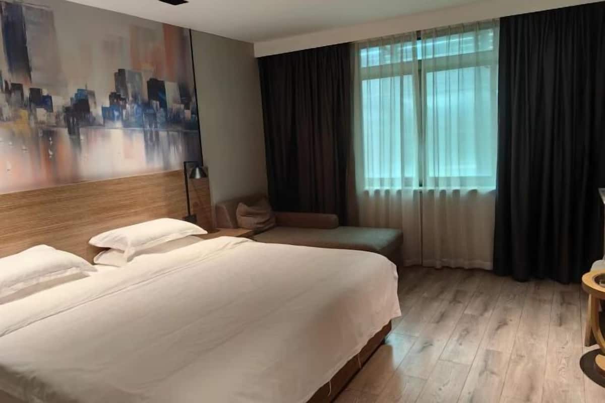 A hotel room with a large bed, a bench, a cityscape painting, and a window with curtains.
