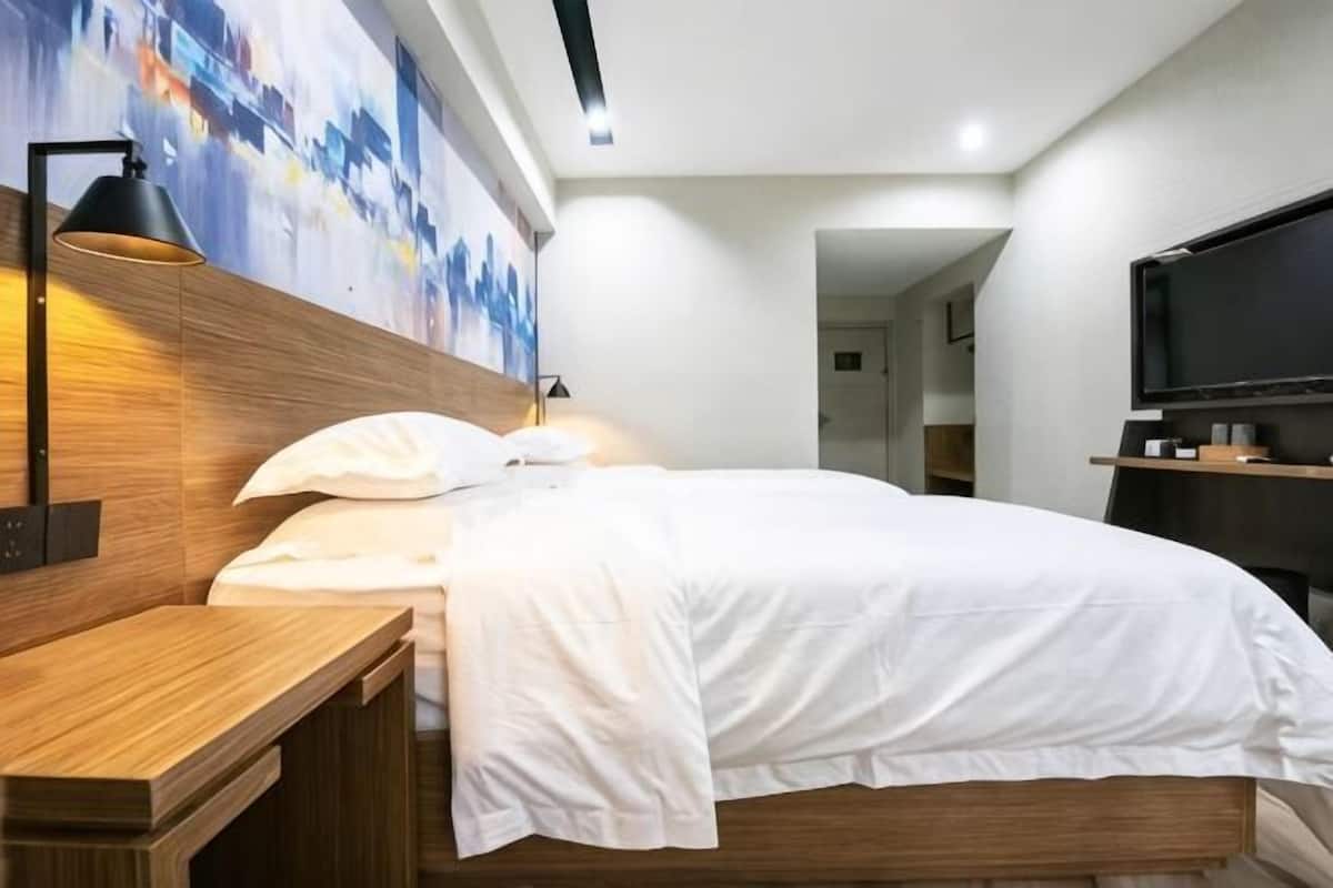 A modern hotel room with a wooden headboard, a bed with white bedding, a nightstand, a wall-mounted TV, and a painting above the bed.