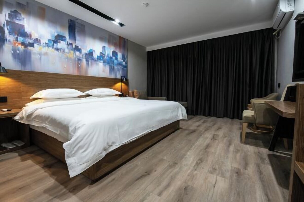 A modern hotel room with a large bed, a desk, and a cityscape painting on the wall.