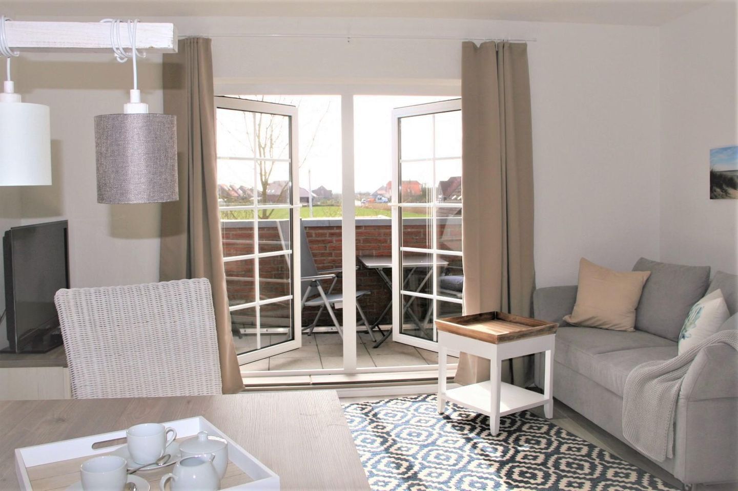 Ein gemütliches Zimmer mit einer Couch, einem Esstisch mit einer Tablett voller Tassen, einem Korbstuhl und Blick auf eine Terrasse mit Tisch und Stühlen.