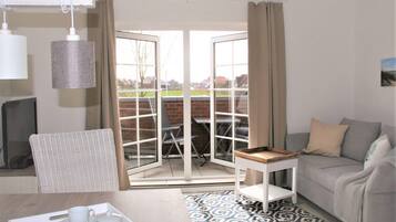 Ein gemütliches Zimmer mit einer Couch, einem Esstisch mit einer Tablett voller Tassen, einem Korbstuhl und Blick auf eine Terrasse mit Tisch und Stühlen.