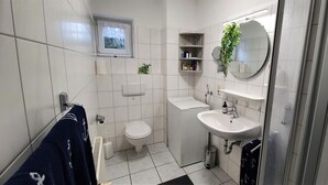 Une salle de bain avec des toilettes, un lavabo et un miroir. Il y a une fenêtre au-dessus des toilettes et une étagère avec des articles de toilette.
