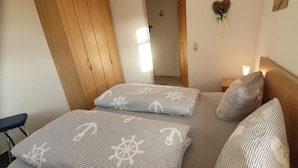 Une chambre avec deux lits, chacun doté d’une literie à rayures et d’une tête de lit. On y trouve une armoire en bois et une petite table de chevet avec une lampe.