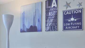 A modern living room with a tufted grey sofa, a round metal coffee table, and three wall art pieces including a sign for 'Caution Low Flying Aircraft'.