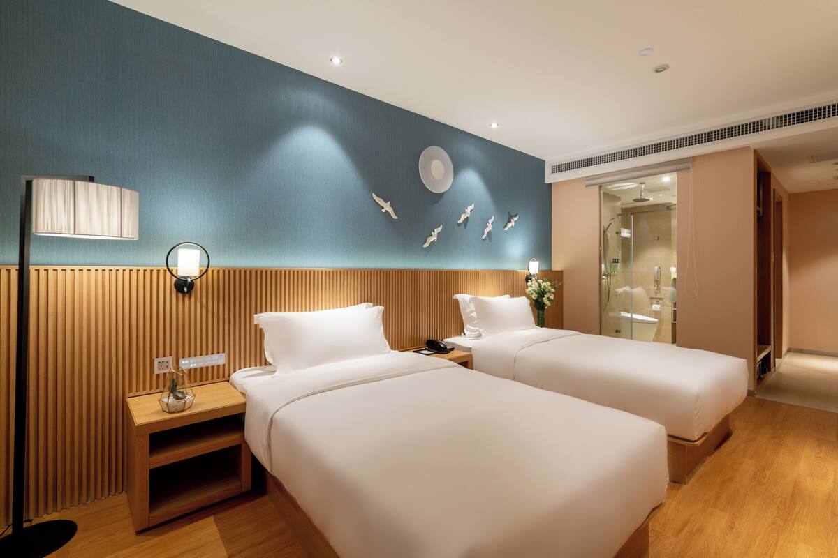 Habitación de hotel con dos camas, cabeceros de madera, una mesita de noche con lámpara y una pared con diseño circular y siluetas de pájaros.