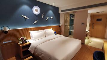 A hotel room with a large bed, a bedside table with a lamp, a wall-mounted phone, and a shower area visible through a glass door.