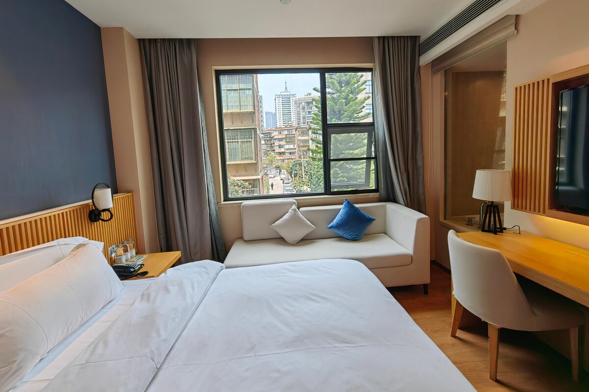 Habitación de hotel con una cama grande, un sofá con cojines, un escritorio con una silla, un televisor y una ventana con vistas a edificios.