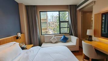 Camera d'albergo con un letto grande, un divano con cuscini, una scrivania con una sedia, una TV e una finestra vista edifici.