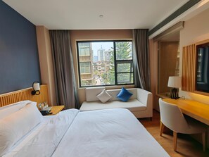 Habitación de hotel con una cama grande, un sofá con cojines, un escritorio con una silla, un televisor y una ventana con vistas a edificios.