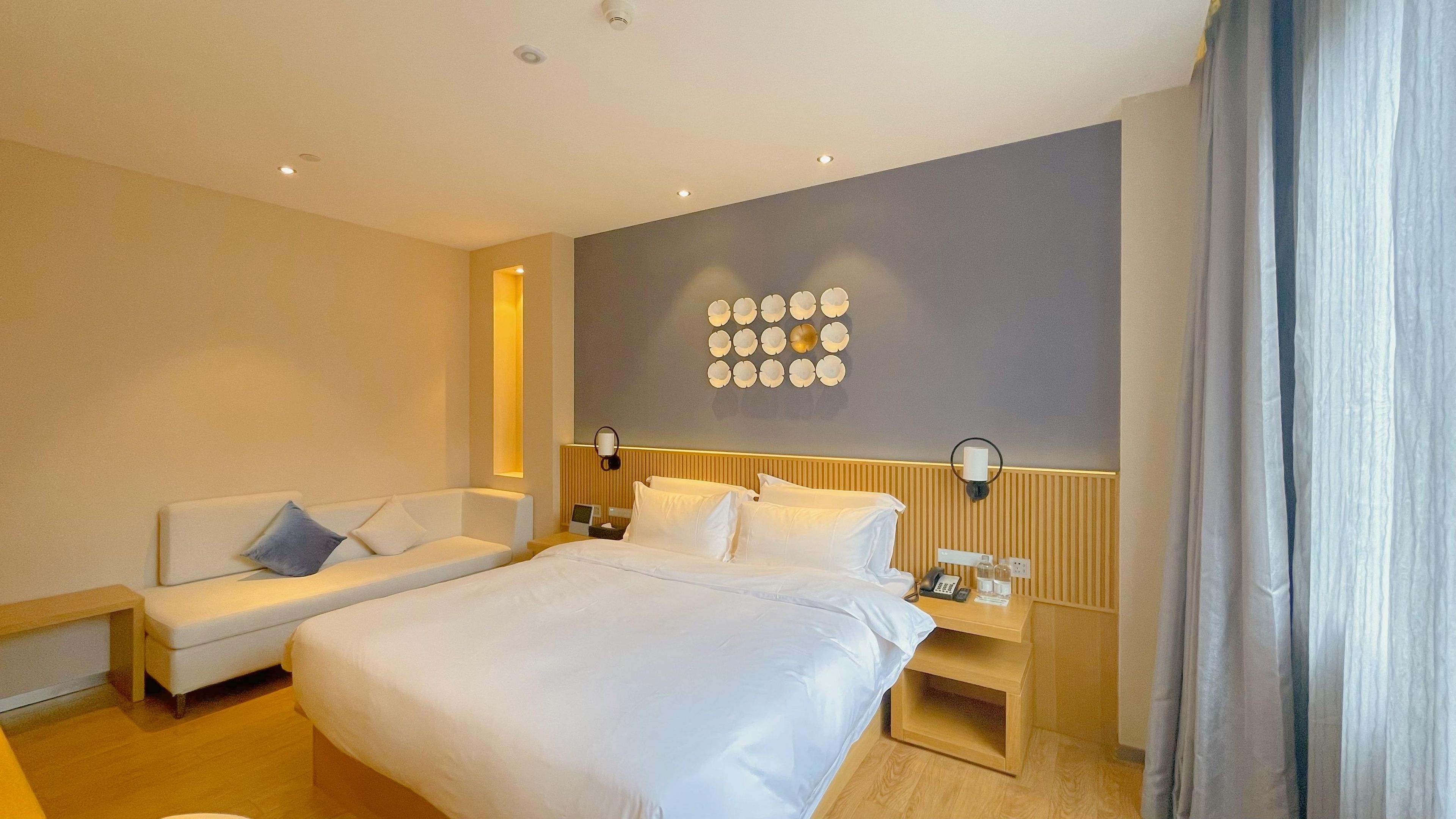 A hotel room with a large bed, a sofa, a bedside table with a lamp, and a wall-mounted light fixture.