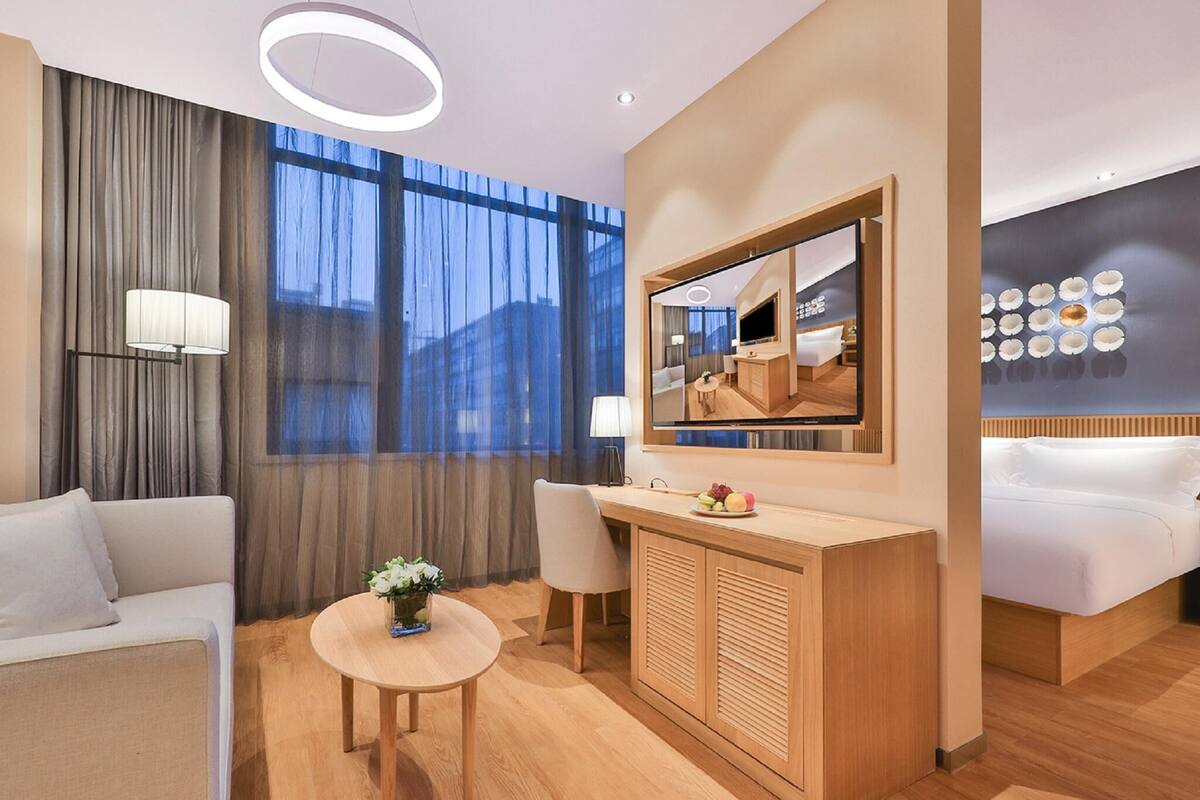 Una habitación de hotel moderna con sofá, una mesa redonda con un florero, minibar, televisión y un amplio ventanal con cortinas.