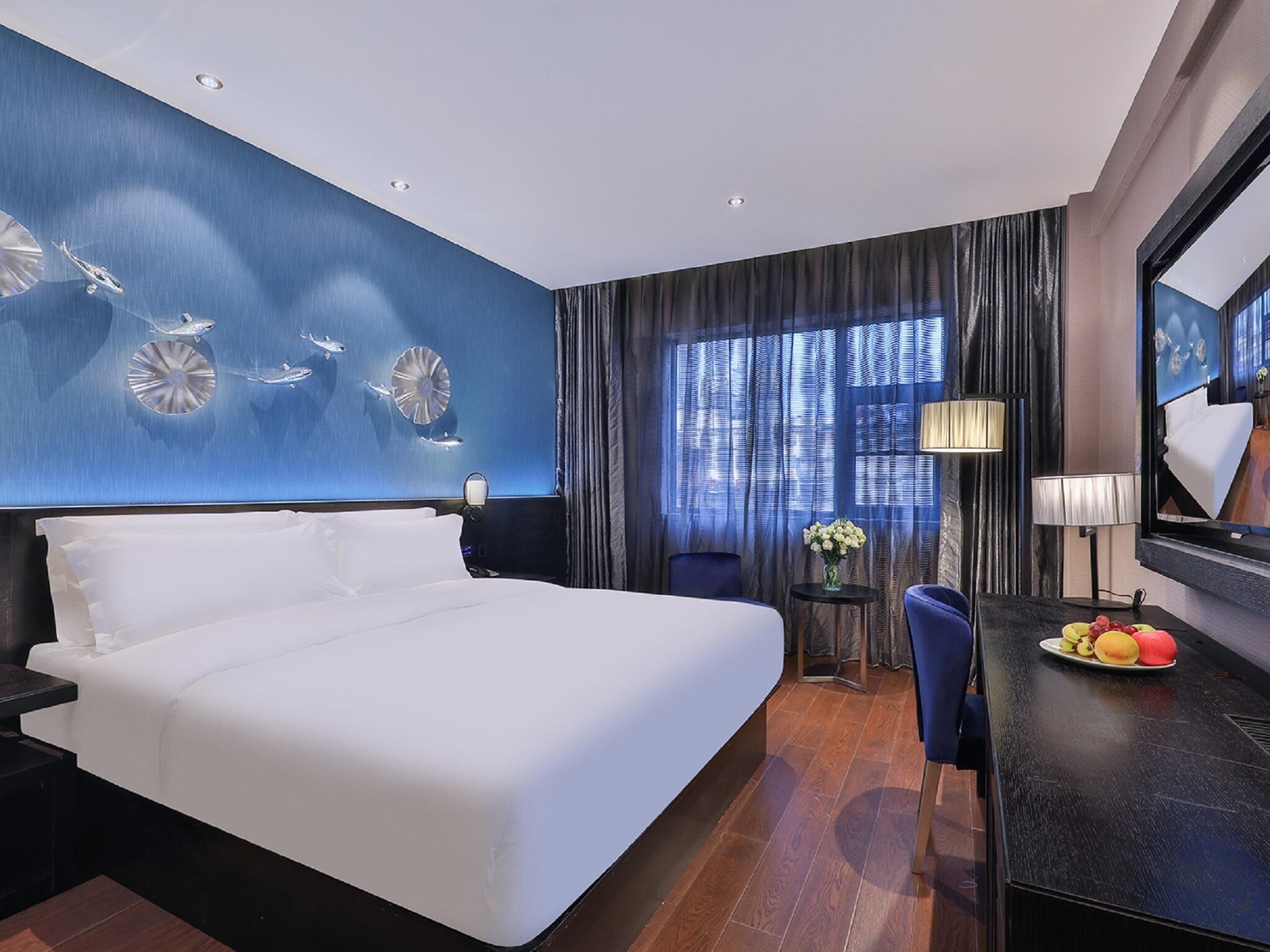 A hotel room with a large bed, a desk with a fruit plate, a chair, a lamp, and a large window with curtains.