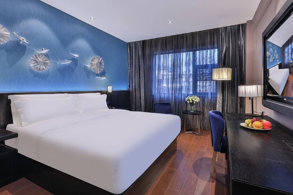Habitación de hotel con una cama grande, un escritorio con una bandeja de frutas, una silla, una lámpara y un ventanal con cortinas.