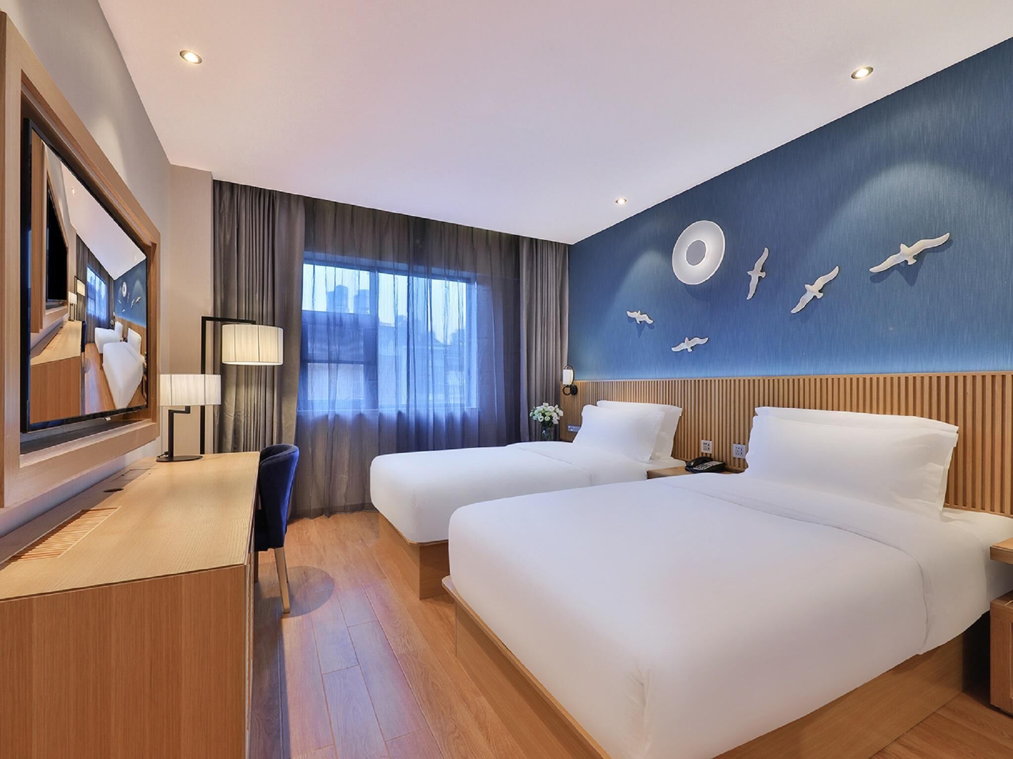 A hotel room with two beds, a desk, a chair, a large mirror, and a wall with a circular light fixture and bird decorations.