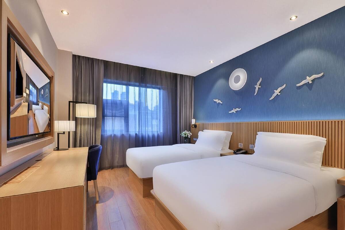 Habitación de hotel con dos camas, un escritorio, una silla, un espejo grande y una pared con lámpara circular y adornos de pájaros.