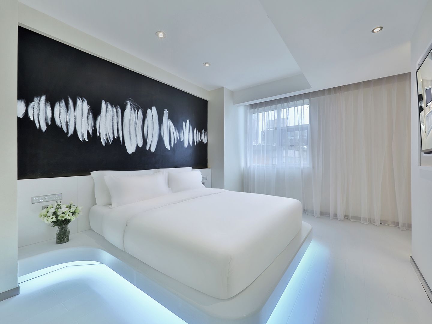 A modern bedroom with a large bed, a black and white abstract painting, and a view of the outdoors through sheer curtains.