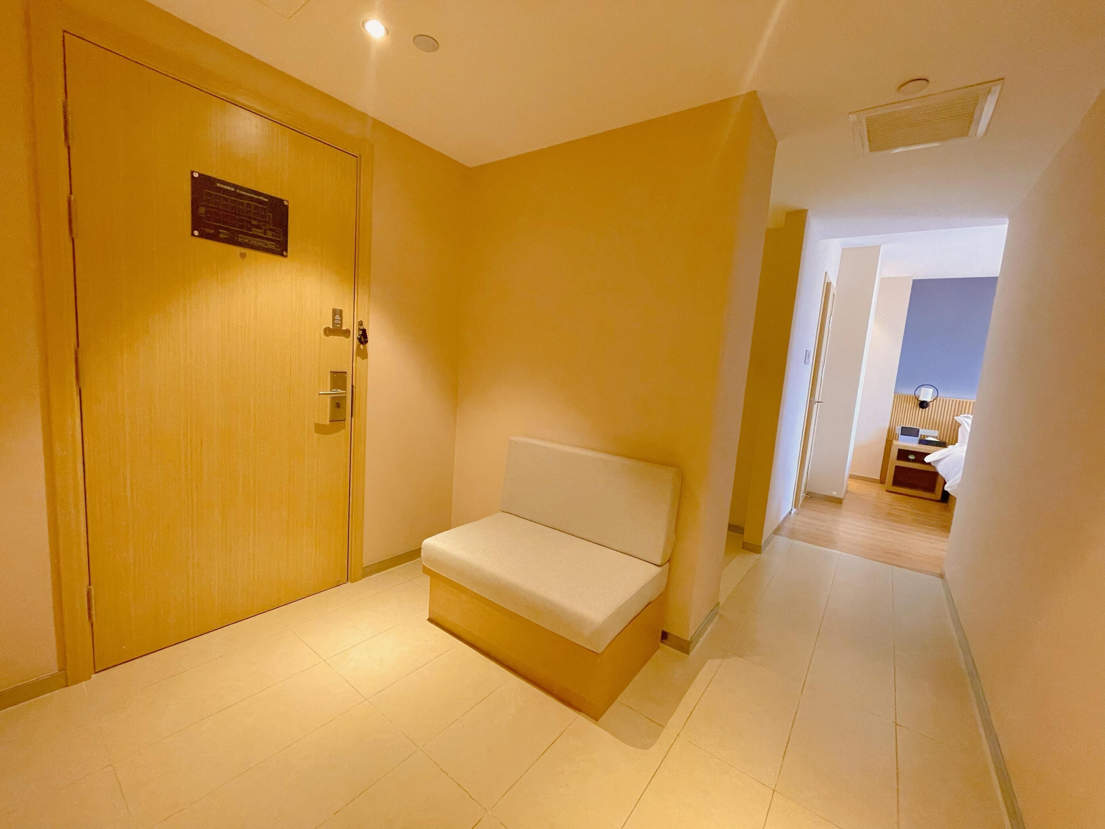 A hotel hallway with a wooden door, a bench, and a bed in the background.