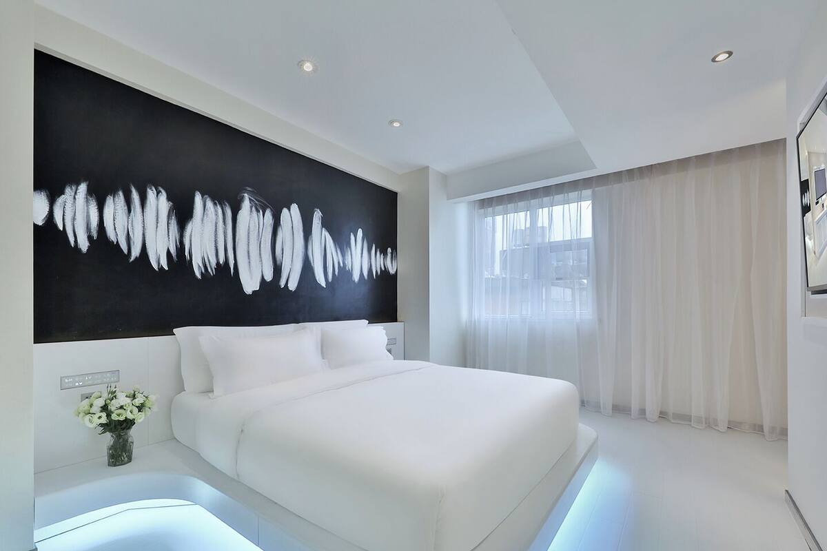 Un dormitorio moderno con una cama grande, un cuadro abstracto en blanco y negro, y vistas al exterior a través de cortinas translúcidas.