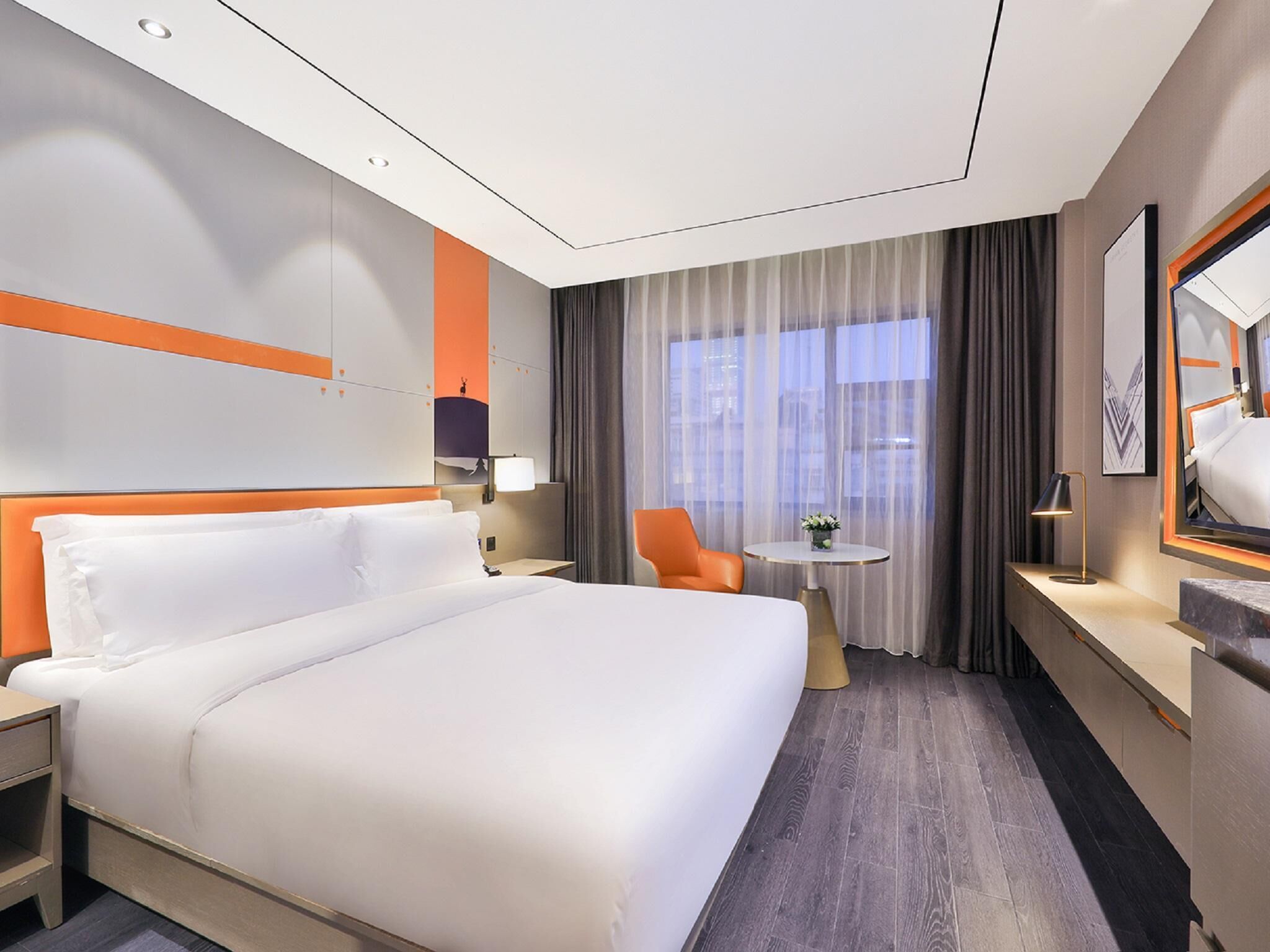 A modern hotel room with a large bed, a desk, a chair, a small table, and a large window with curtains.