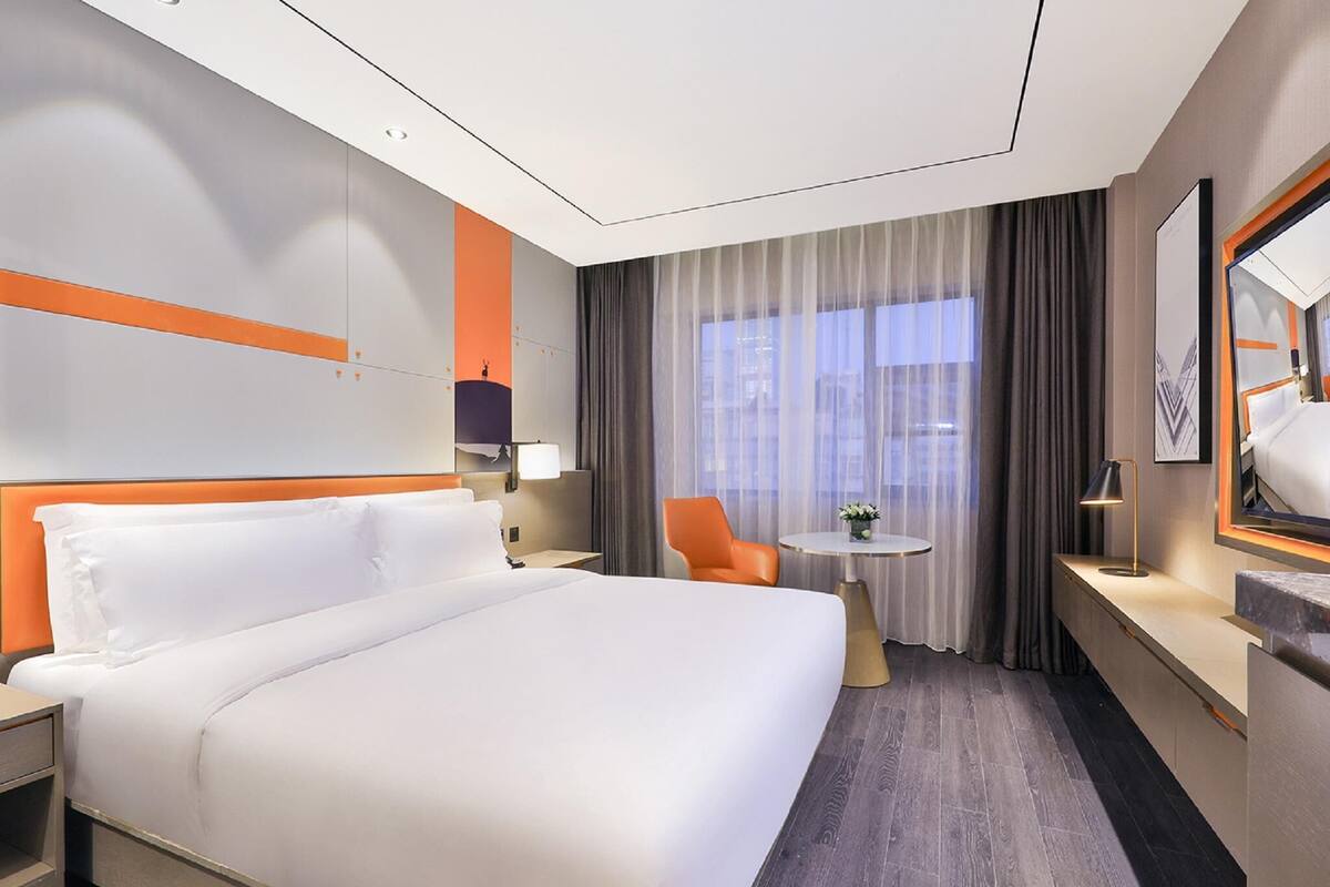 Una habitación de hotel moderna con una cama grande, un escritorio, una silla, una mesita y un ventanal con cortinas.