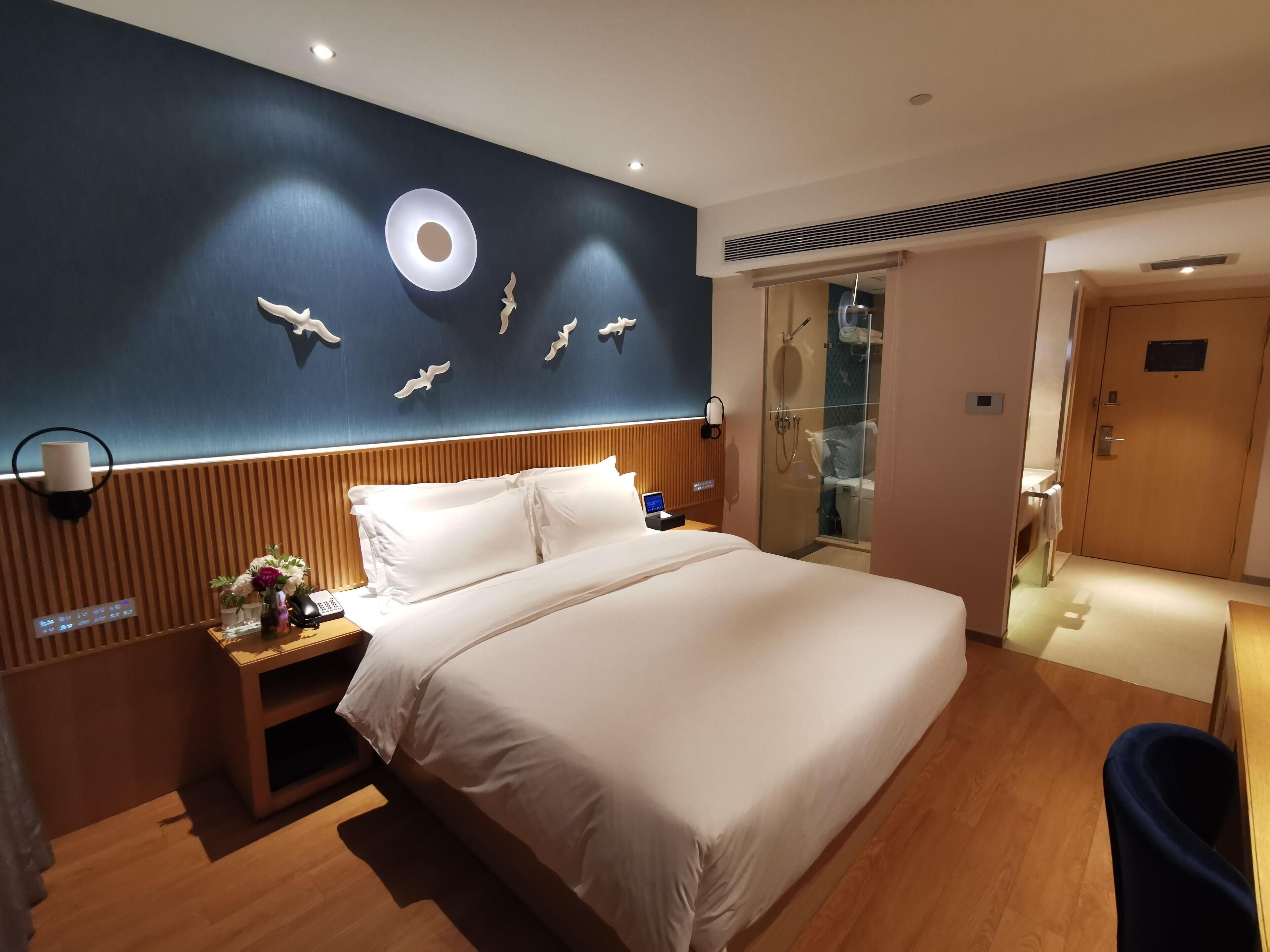 A hotel room with a large bed, a bedside table with a lamp, a wall-mounted phone, and a shower area visible through a glass door.