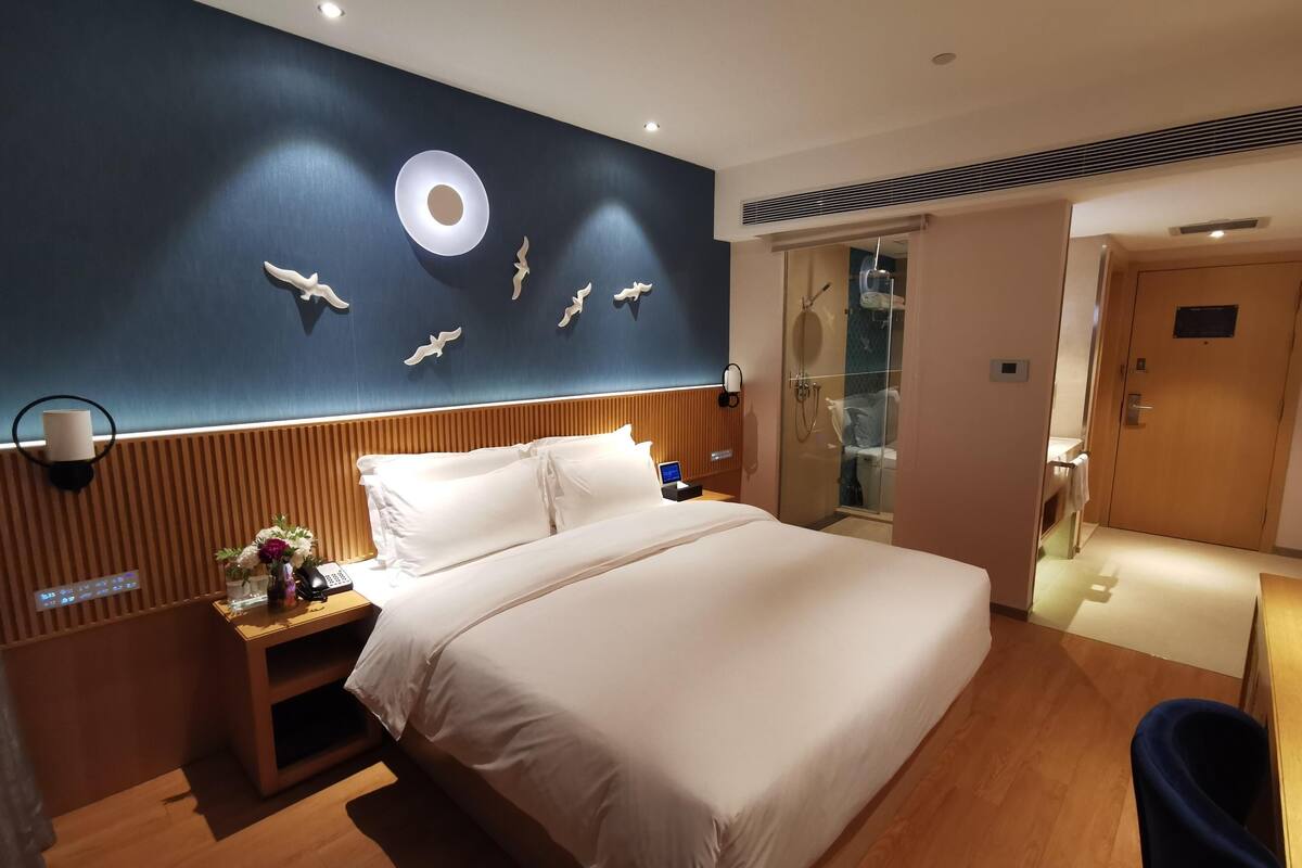 Habitación de hotel con una cama grande, una mesita de noche con lámpara, teléfono en la pared y zona de ducha visible a través de un vidrio.