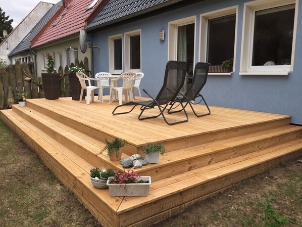 Eine Holzterrasse mit Sitzgelegenheiten im Freien, Blumentöpfen und Blick auf ein Haus mit blauer Fassade und rotem Dach.
