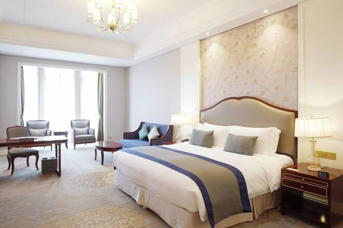 A hotel room with a large bed, a desk, two chairs, a chandelier, and a large window.